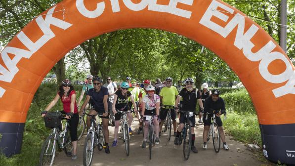 A group of cyclists are in a line about to set off. They are under a big rubber archway that reads 'walk, cycle, enjoy'